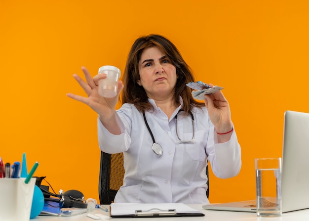 Unpleased middle-aged female doctor wearing medical robe and stethoscope sitting at desk with medical tools laptop and clipboard stretching out medical drugs and beaker isolated