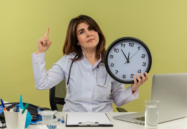 Unpleased middle-aged female doctor wearing medical robe and stethoscope sitting at desk with medical tools and laptop holding clock pointing up isolated