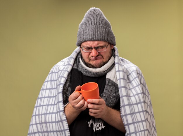 Unpleased middle-aged ill male wearing winter hat and scarf wrapped in plaid holding and looking at cup of tea 