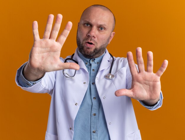 Unpleased middle-aged male doctor wearing medical robe and stethoscope  doing stop gesture isolated on orange wall
