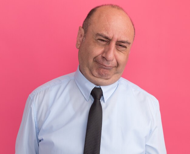 Недовольный мужчина средних лет в белой футболке с галстуком на розовой стене