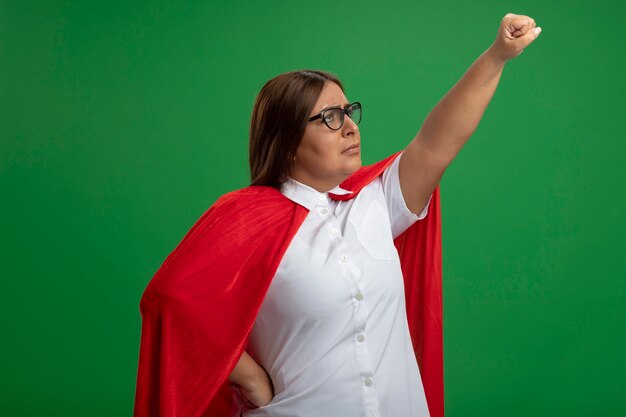 Unpleased middle-aged superhero female raising fist wearing glasses isolated on green