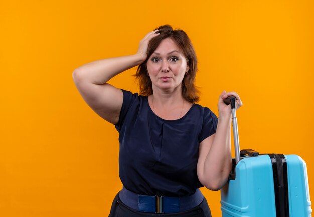 Unpleased middle-aged traveler woman holding suitcase and putting hand on head on isolated orange