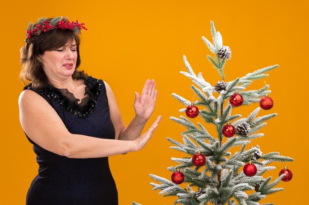 Unpleased middle-aged woman wearing christmas head wreath and tinsel garland around neck standing near decorated christmas tree