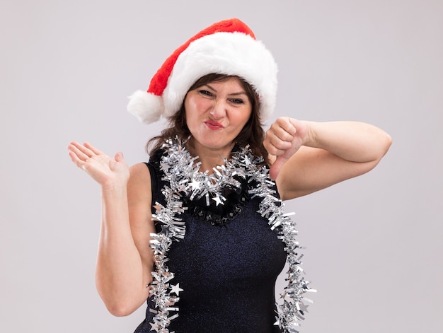 Недовольная женщина средних лет в новогодней шапке и мишурной гирлянде на шее, глядя в камеру, показывая пустую руку и большой палец вниз, изолированные на белом фоне