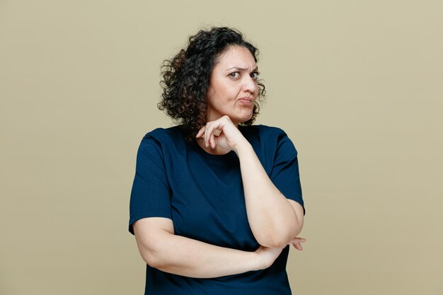 Unpleased middleaged woman wearing tshirt keeping hand under elbow and another hand near chin looking at camera isolated on olive green background