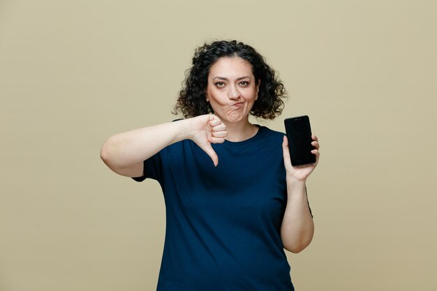 недовольная женщина средних лет в футболке с мобильным телефоном, смотрящая в камеру, показывает большой палец вниз на оливково-зеленом фоне