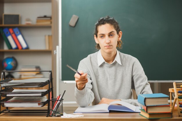 unpleased points atside with pointer young male teacher sitting at desk with school tools on in classroom