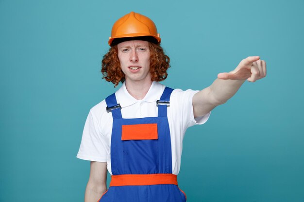 Unpleased points at side young builder man in uniform isolated on blue background