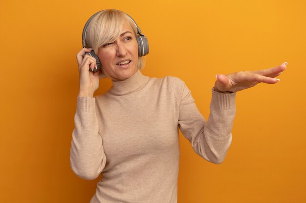 Unpleased pretty blonde slavic woman on headphones looks and points at side with hand on orange