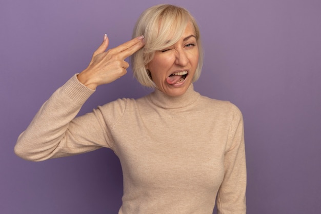 Free photo unpleased pretty blonde slavic woman stucks out tongue and gestures gun hand sign putting at temple on purple