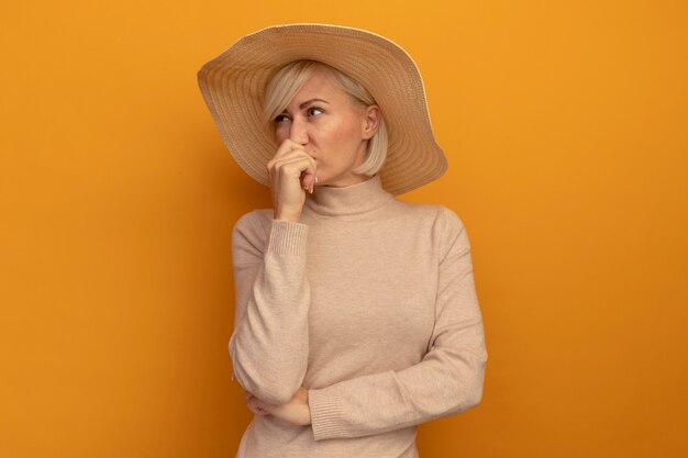 Unpleased pretty blonde slavic woman with beach hat holds hand in front of mouth isolated on orange wall