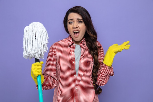 Unpleased pretty caucasian cleaner woman with rubber gloves holding mop and keeping hand open 