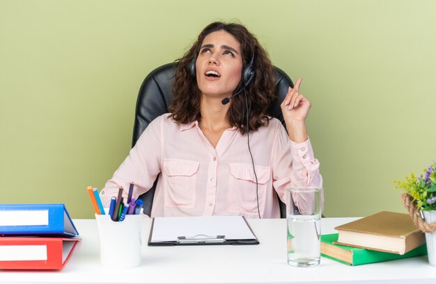 Unpleased pretty caucasian female call center operator on headphones sitting at desk with office tools looking and pointing up isolated on green wall