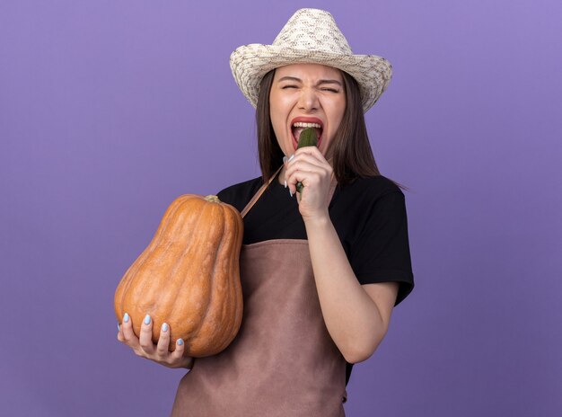 Unpleased pretty caucasian female gardener wearing gardening hat holding pumpkin and biting cucumber isolated on purple wall with copy space