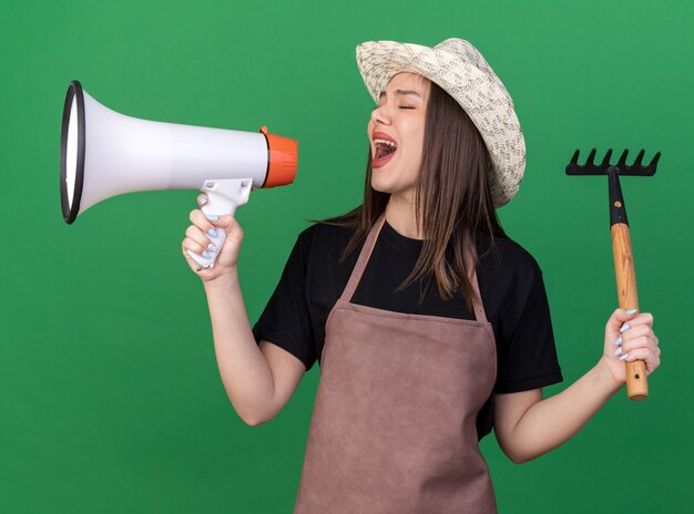 Unpleased pretty caucasian female gardener wearing gardening hat holding rake and shouting into loud speaker