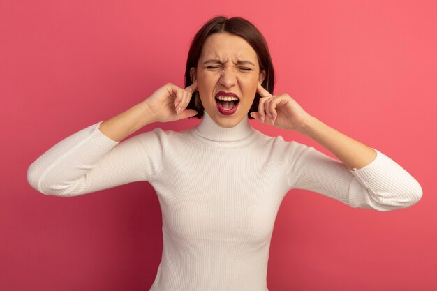 Unpleased pretty caucasian woman closes ears with fingers on pink