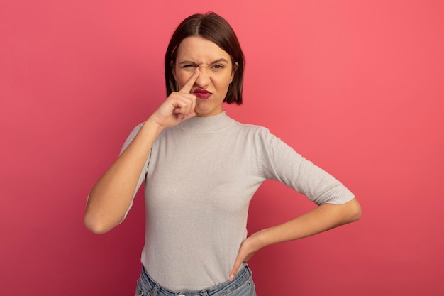 Unpleased pretty caucasian woman puts finger on nose on pink