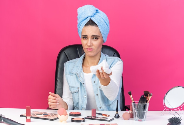Unpleased pretty caucasian woman with wrapped hair in towel sitting at table with makeup tools holding out insert napkin 