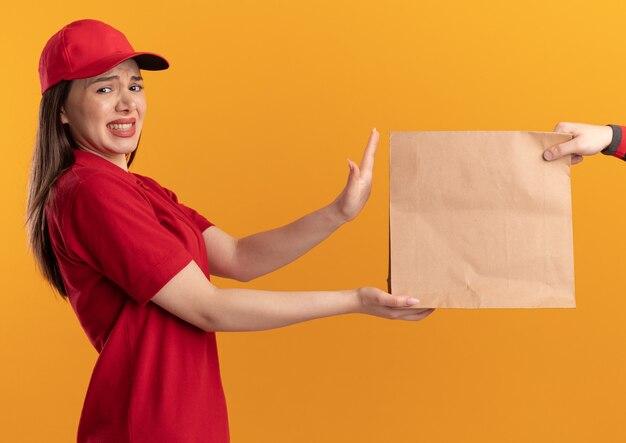 Unpleased pretty delivery woman in uniform gives paper package to someone looking at camera isolated on orange wall with copy space