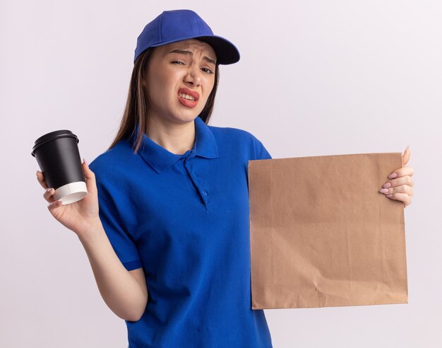 Unpleased pretty delivery woman in uniform holds paper package and paper cup on white