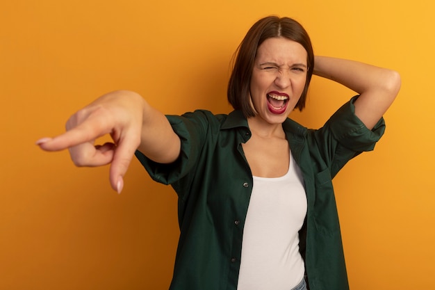 Unpleased pretty woman puts hand on head behind looking and pointing at side isolated on orange wall