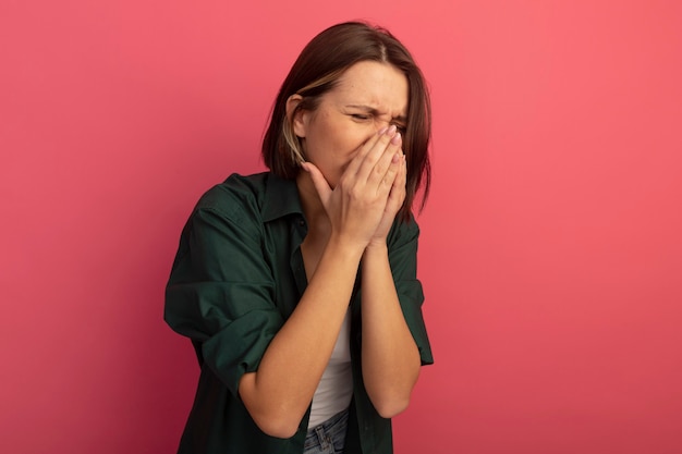 Unpleased pretty woman puts hands on nose isolated on pink wall