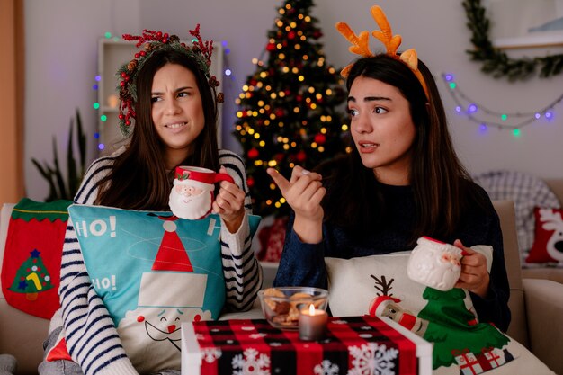 Недовольные симпатичные молодые девушки с венком из падуба и ободком с оленями держат чашки и смотрят в сторону, сидя на креслах и наслаждаясь Рождеством дома