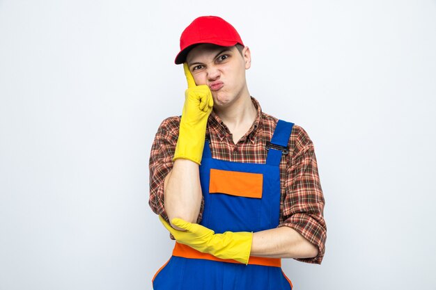 Unpleased putting finger on cheek young cleaning guy wearing uniform and cap with gloves 