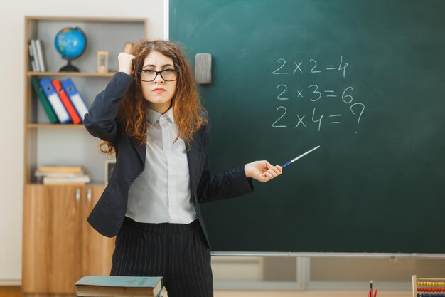 unpleased putting hand on head young female teacher holding pointer standing in front blackboard in classroom