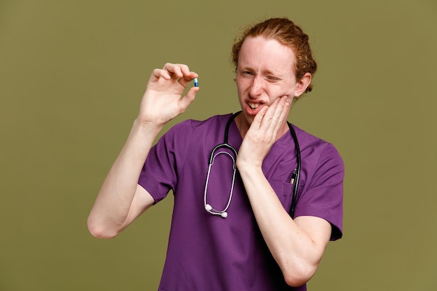 Бесплатное фото Недовольный кладет руку на щеку с таблетками молодой врач-мужчина в униформе со стетоскопом на зеленом фоне
