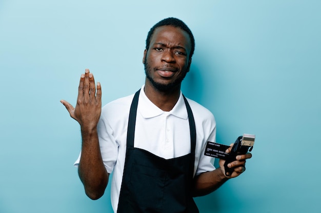 Unpleased raised hand holding card with hair clippers young african american barber in uniform isolated on blue background