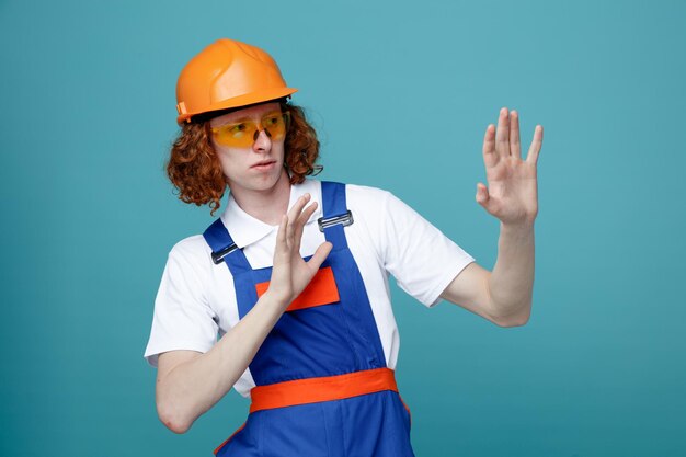 Scontento alzando gand giovane costruttore uomo in uniforme isolato su sfondo blu