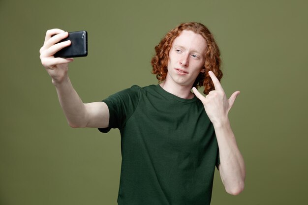 unpleased showing goat gesture young handsome guy wearing green t shirt take a selfie isolated on green background