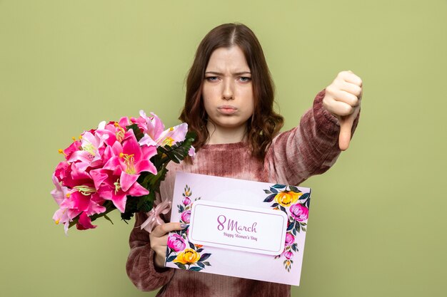 オリーブグリーンの壁に分離されたはがきと花束を保持している幸せな女性の日に美しい少女を親指で示すのは不愉快です