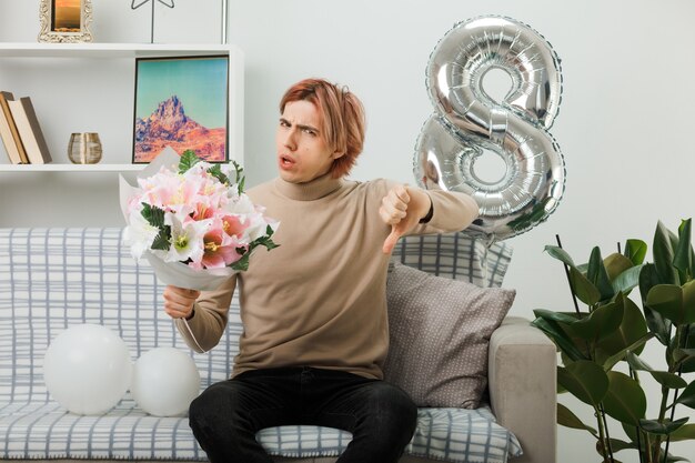 Unpleased showing thumb down handsome guy on happy women day holding bouquet sitting on sofa in living room