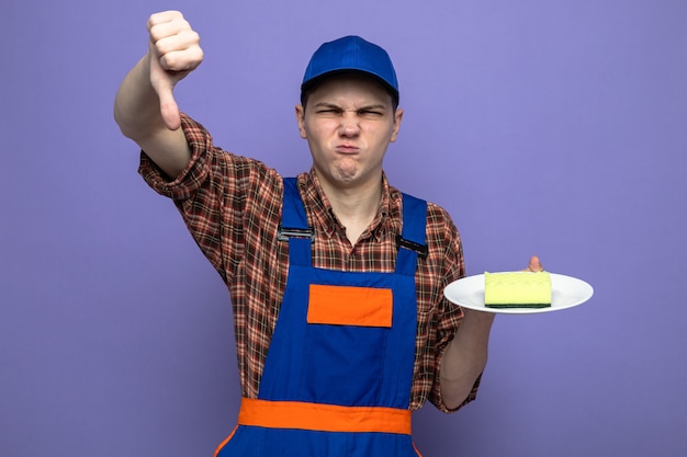 Free photo unpleased showing thumb down young cleaning guy wearing uniform and cap holding sponge on plate
