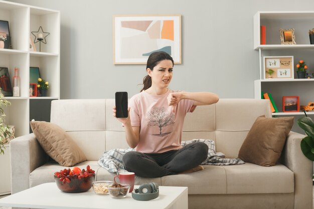 Unpleased showing thumb down young girl holding phone sitting on sofa behind coffee table in living room