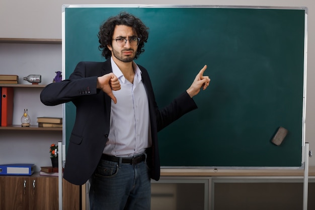 Unpleased showing thumb down young male teacher wearing glasses points at blackboard in classroom