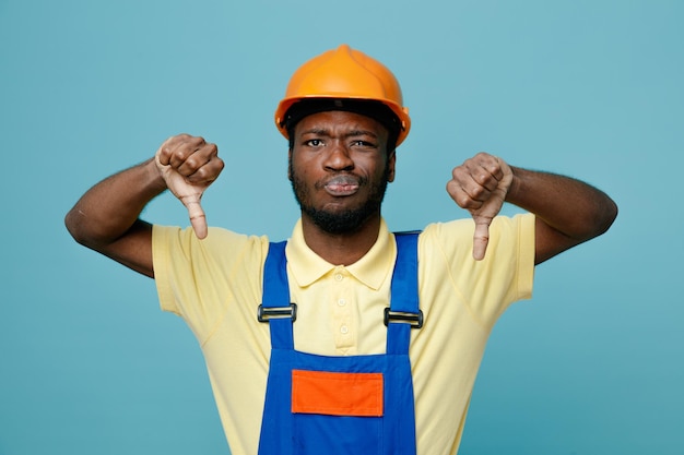 Недовольный показывает палец вниз молодому афро-американскому строителю в униформе, изолированному на синем фоне