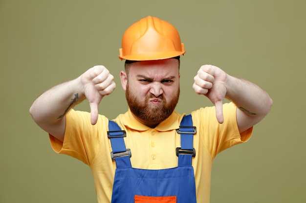 Unpleased showing thumbs down young builder man in uniform isolated on green background