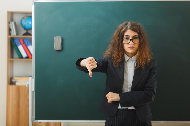 교실에서 칠판 앞에 서 있는 젊은 여성 교사에게 엄지손가락을 아래로 보여주는 불쾌한