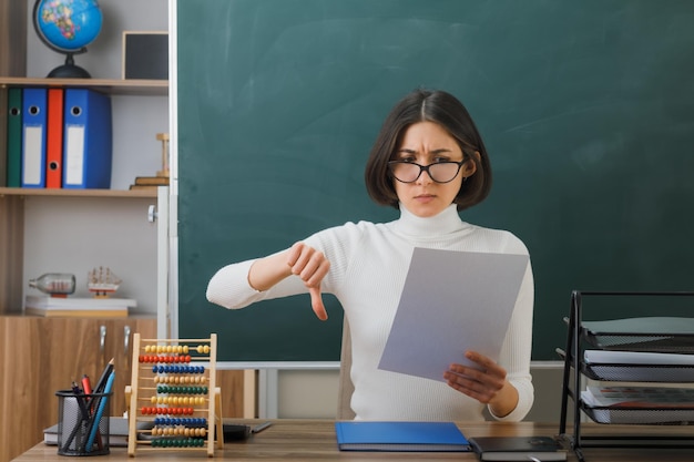 unpleased showing thumbs down young female teacher wearing glasses holding paper sitting at desk with school tools on in classroom