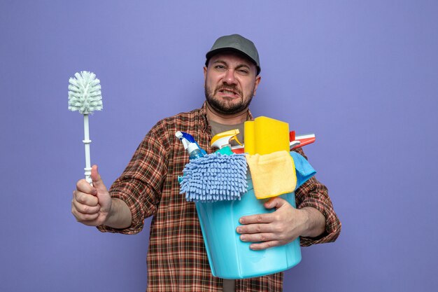 Unpleased slavic cleaner man holding cleaning equipment and toilet brush