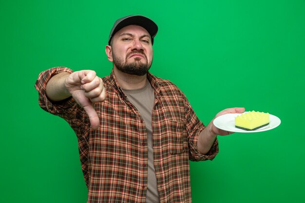 Unpleased slavic cleaner man holding sponge on plate and thumbing down 