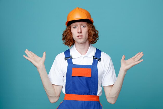 Недовольный разводящий руки молодой строитель в форме, изолированный на синем фоне