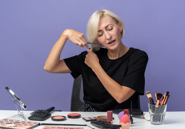 Unpleased with closed eyes young beautiful girl sits at table with makeup tools cutting hair with scissors isolated on blue background