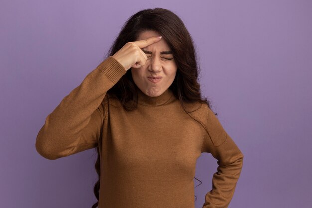 Unpleased with closed eyes young beautiful girl wearing brown turtleneck sweater putting finger on forehead isolated on purple wall