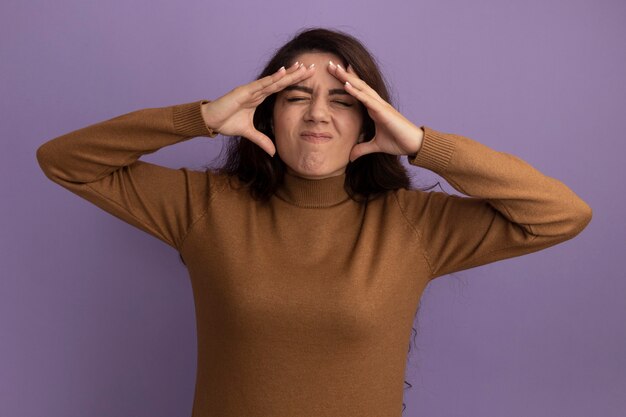 Unpleased with closed eyes young beautiful girl wearing brown turtleneck sweater putting hands on temple isolated on purple wall