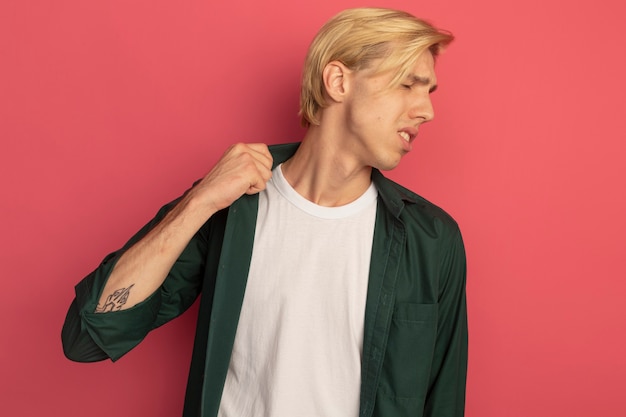 Unpleased with closed eyes young blonde guy wearing green t-shirt holding collar isolated on pink